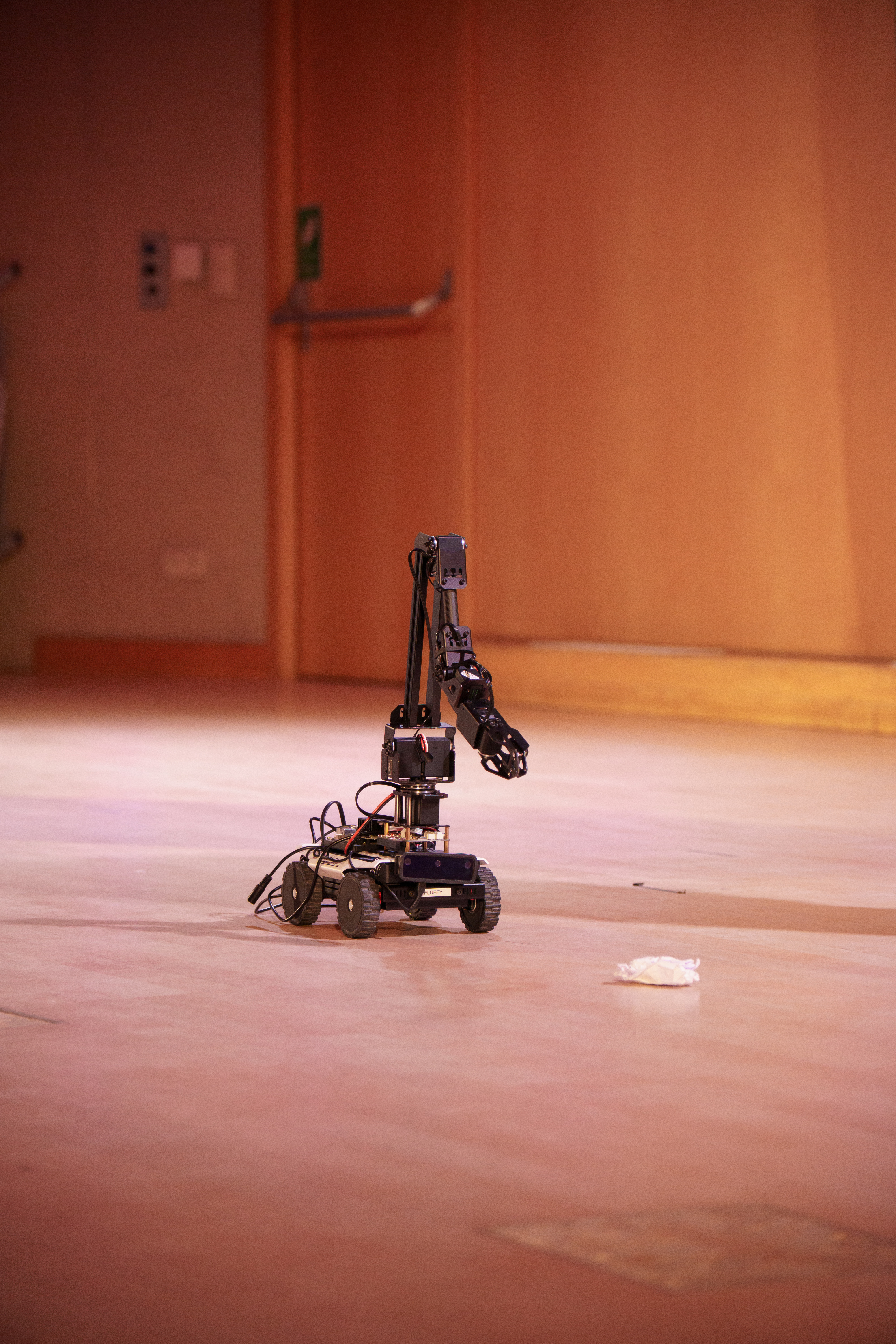 GHOST's riverside cleanup robot extends its black manipulator arm toward a crumpled piece of litter on a hard floor, in a wide empty hall.